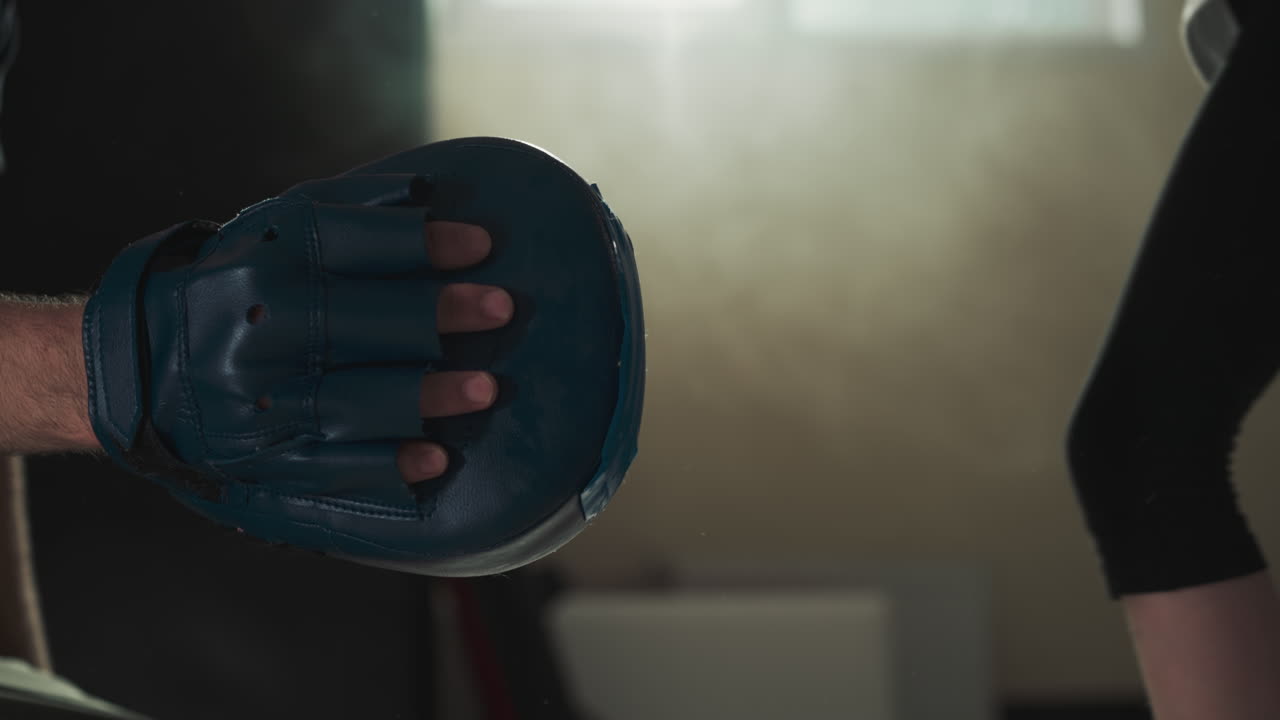 Child kicks pad by leg with trainer in gym closeup. Combat instructor conducts training with little girl in sports school slow motion. Children hobby