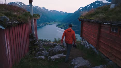 Man-walking-in-between-two-small-and-old-houses-towards-a-rock,-watching-over-a-beautiful-landscape-of-river-and-mountain-coming-together