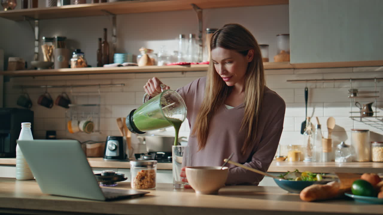 Girl watching video recipe at laptop preparing vegetables smoothie in kitchen.
