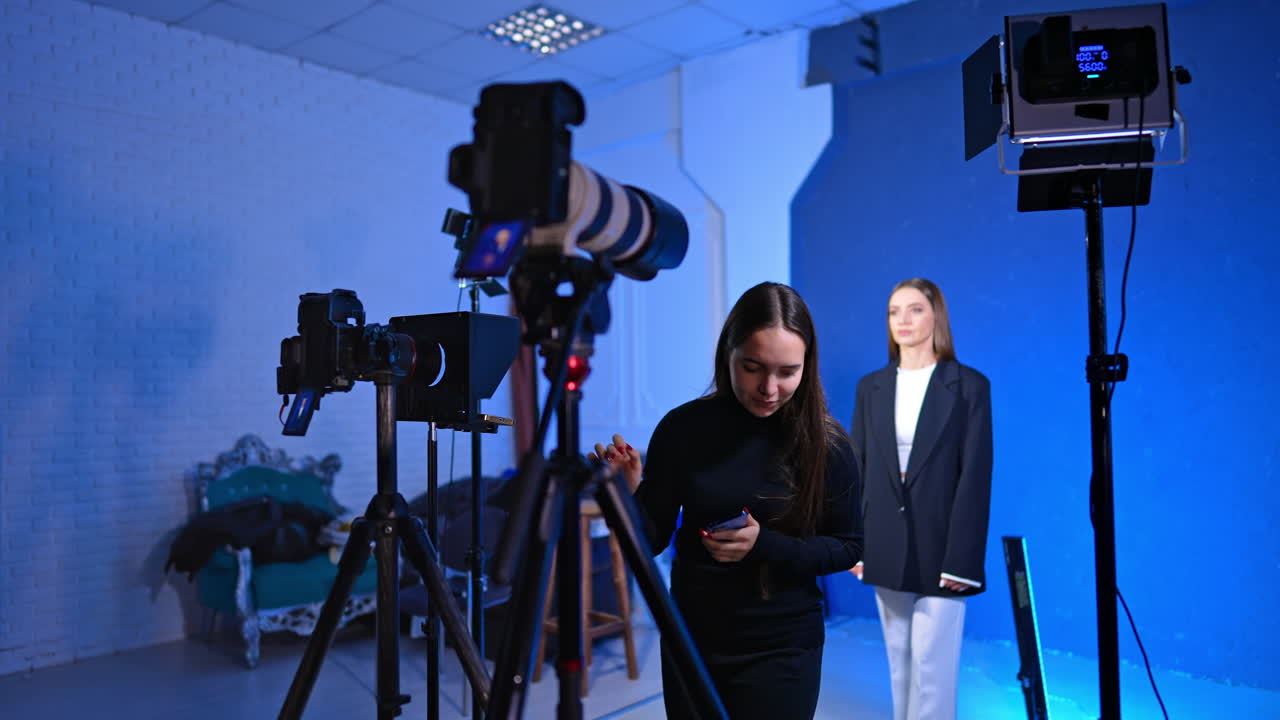 Asistente revisando la configuración de la cámara de video durante una sesión de estudio profesional. Una mujer con jersey de cuello alto negro está junto al equipo de cámara con un presentador en traje blanco de fondo contra una pared azul.