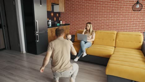 father plays with his child at home while their mother busy with her laptop on the sofa