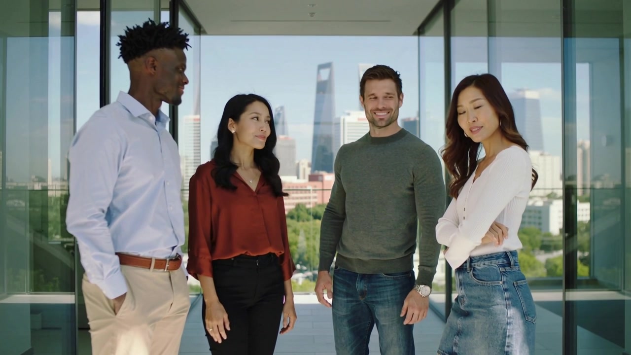 Un team di quattro persone, appoggiato e che parla, condivide aggiornamenti su un balcone di vetro, in abiti eleganti, con lo skyline.