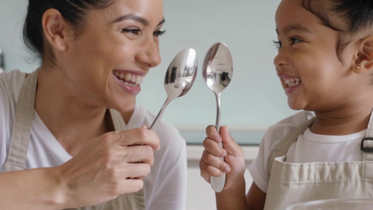 Madre e figlia con grembiuli beige che alzano i cucchiai, facendoli tintinnare e creando un legame in cucina, con una ciotola di farina.