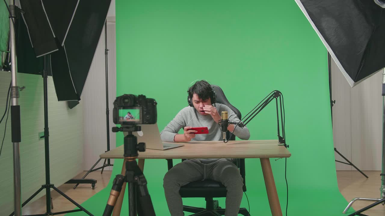 A Camera Recording Asian Man Gamer With Headphone And Computer Losing Mobile Phone Game On The Green Screen Background With Professional Light Equipment