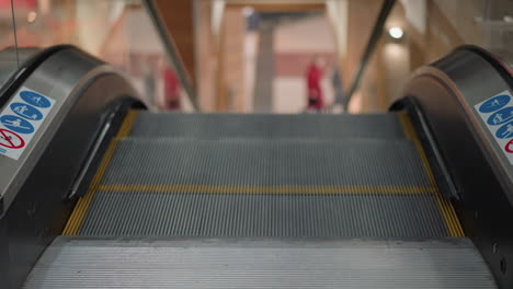 entrance view of moving walkway ascending through glass balustrade in busy mall environment, blurred figures in background convey motion, metallic steps with sleek handrails, public transport element