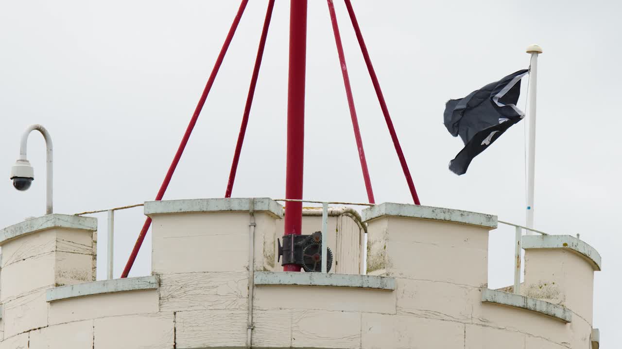 Black flag waves vigorously atop stone tower with red cables, overcast daylight, static camera