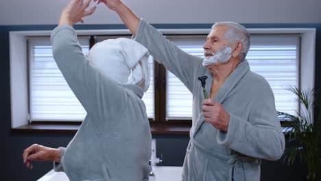 Senior-couple-man-and-woman-in-bathrobe-are-dancing-in-bathroom-having-fun-enjoying-leisure-time