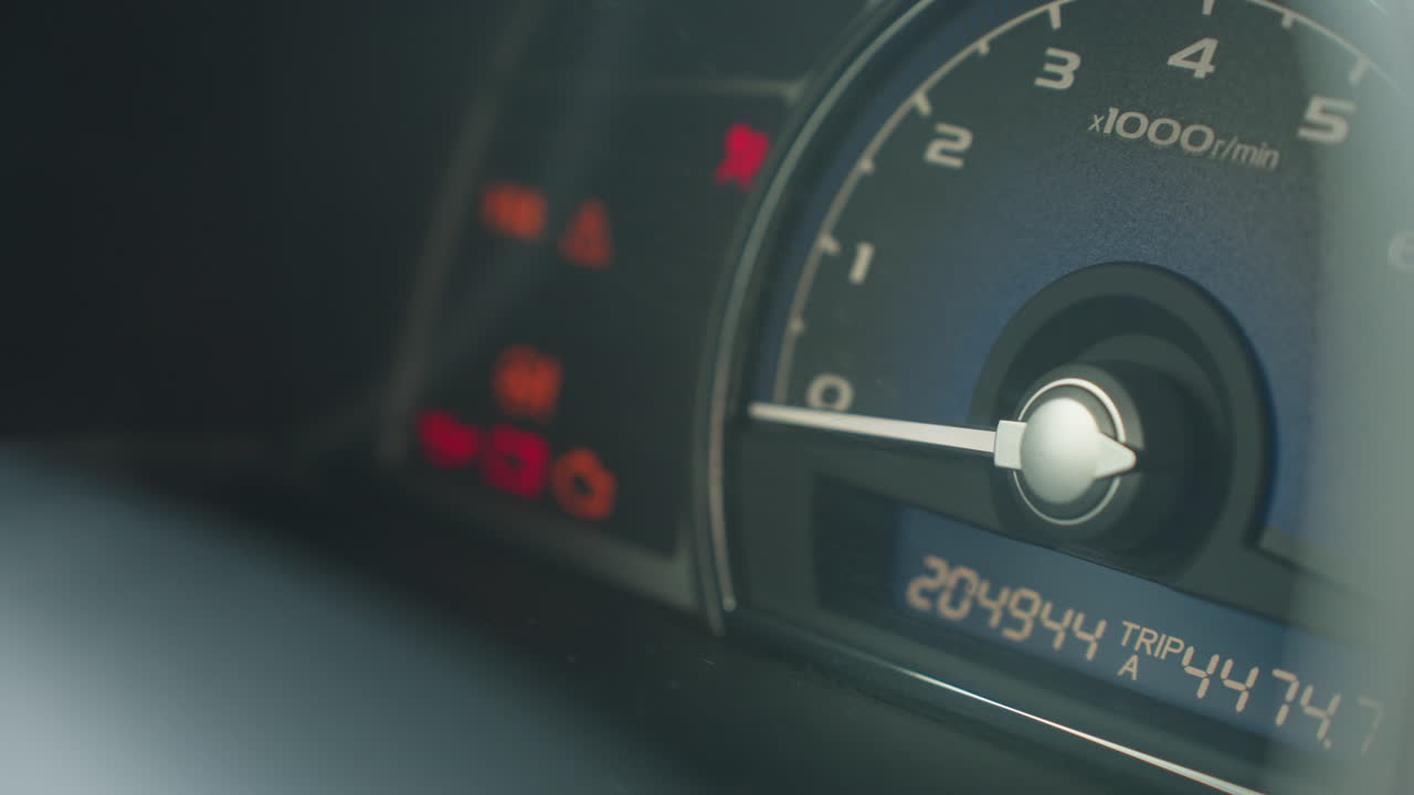 close up of illuminated tachometer gauge in running vehicle interior, needle resting above zero rpm, glowing dial markings visible through steering wheel blur, engine on indicator active