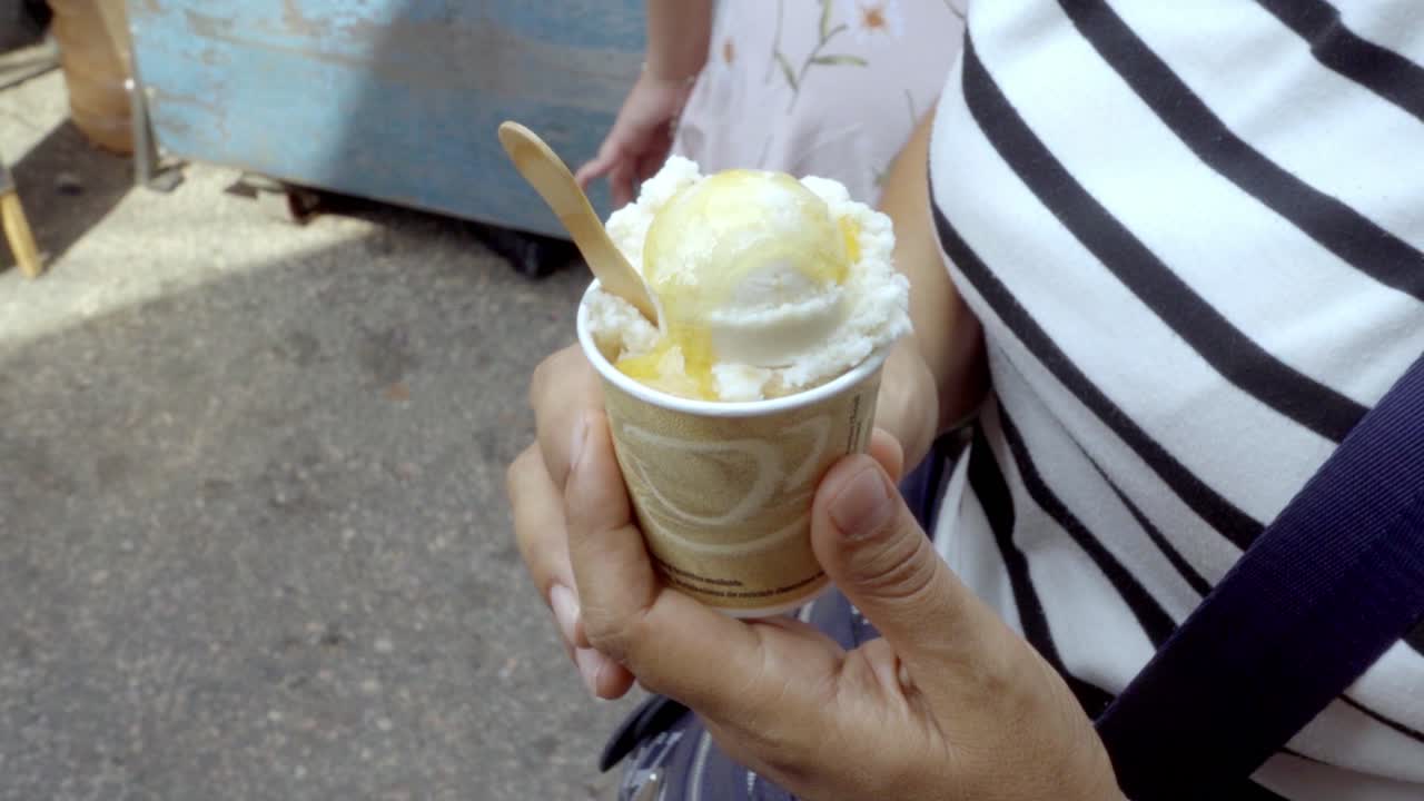 Primo piano di una persona che tiene un bicchiere di carta riempito con gelato bianco, guarnito con miele dorato e un cucchiaino di legno. Perfetto per contenuti di viaggio estivi e gastronomici.