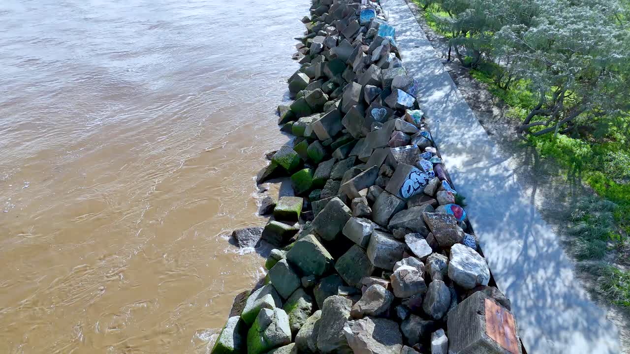 Drone camera glides above a concrete breakwater lined with painted rocks, separating a river from a walking path bordered by greenery in bright daylight