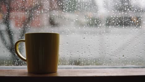 Ein-Dampfender-Gelber-Becher-Voller-Regentropfen-Neben-Dem-Fenster