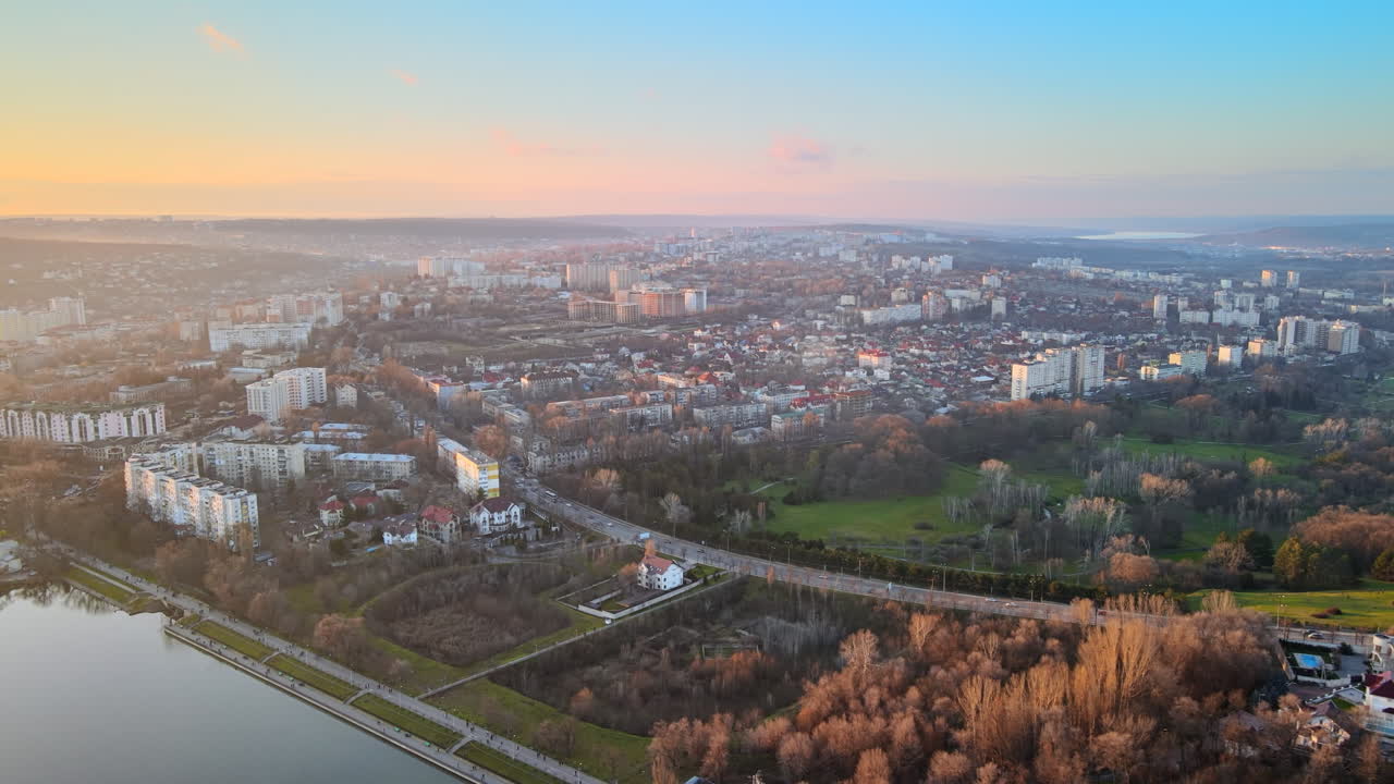 Aerial drone view of Chisinau at sunset. Panorama view of multiple residential buildings, road with cars, bare trees, two parks and lake. Moldova
