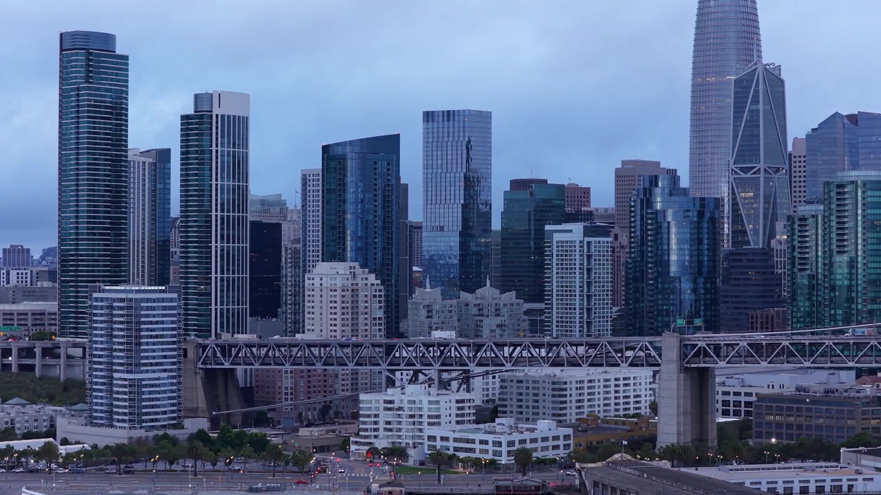 Riprese aeree catturano lo skyline dell'Embarcadero di San Francisco e il Bay Bridge al tramonto, con iconici grattacieli moderni, una calma luce blu serale e un dinamico paesaggio urbano.