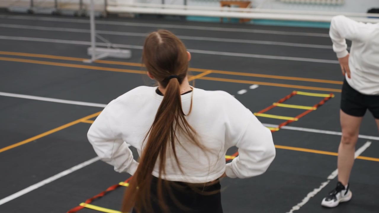 Indoor badminton agility ladder drill coach instructing trainee during focused warmup, quick footwork, teamwork on marked court, sneakers tapping rungs, instructor cues and intensity