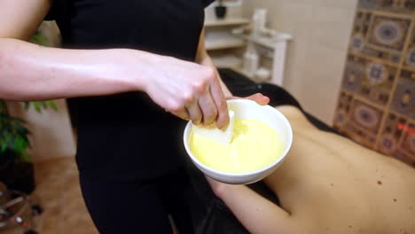 unrecognized female in black clothes mixes the cream in the bowl. massagist applies cream with wide brush on the female back.