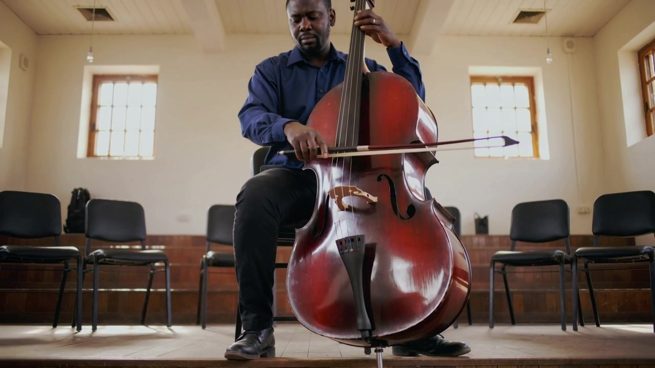 Bringing bow forward, seated navy shirt musician bowing and fingering cello on stage, rehearsing