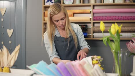 Female-blonde-florist-arranging-modern-bouquet-and-finishing-it-with-perfect-riband.-Handsome-flower-shot-owner-working-at-her