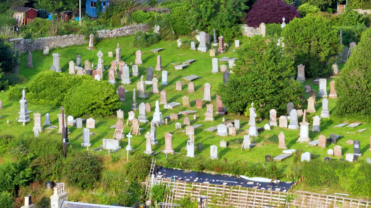 Drone camera smoothly pans above old gravestones, lush greenery, and rural village rooftops in daylight