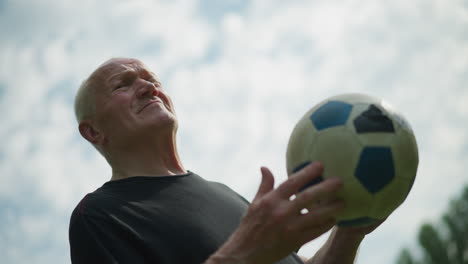 an elderly man holds a soccer ball in his hand, rotating it thoughtfully as he looks around under a cloudy sky