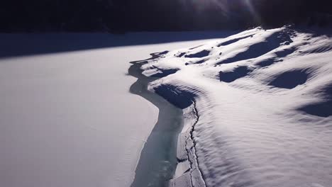 Aerial-view-over-the-shore-of-a-melting-frozen-lake-to-a-huge-mountain-panorama-with-big-snowy-mountains