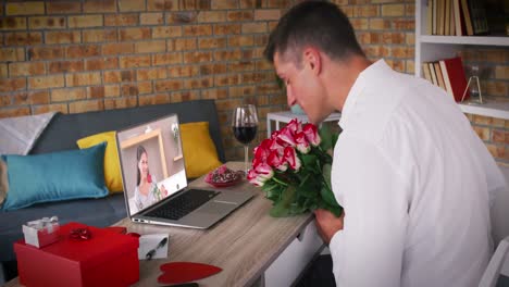 Kaukasisches-Paar-Bei-Einem-Valentinstag-Videoanruf,-Mann-Hält-Blumen-Und-Bläst-Einer-Frau-Auf-Dem-Laptop-Einen-Kuss-Zu