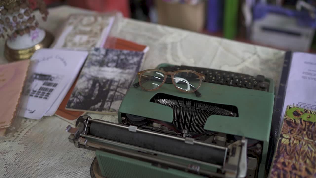 Close-up of a vintage green typewriter with eyeglasses resting on top, surrounded by papers and notebooks on a lace-covered table, evoking a nostalgic writing atmosphere - parallax shot