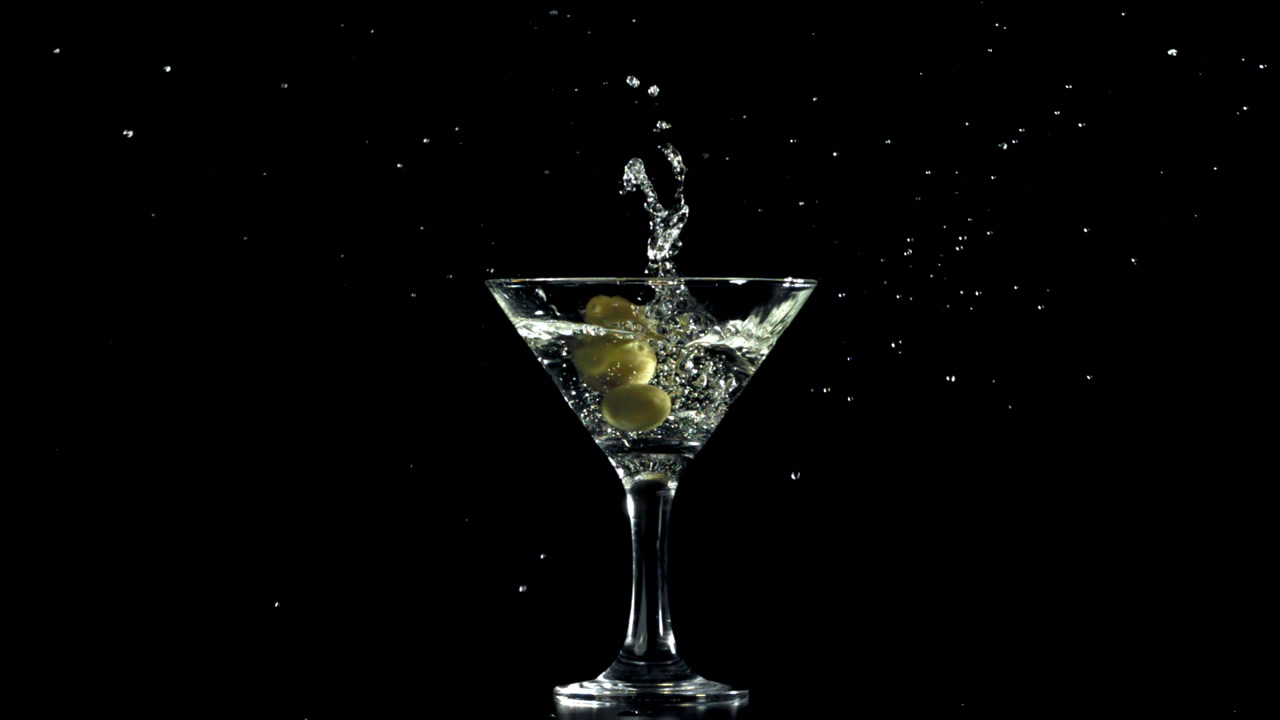Due olive verdi che si tuffano in un bicchiere da Martini al rallentatore