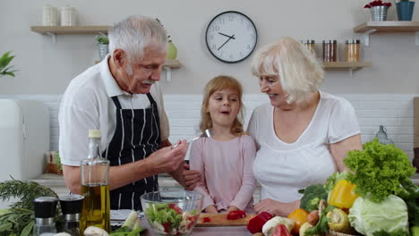 elderly grandparents in kitchen feeding grandchild girl with chopped red pepper. vegetarian diet