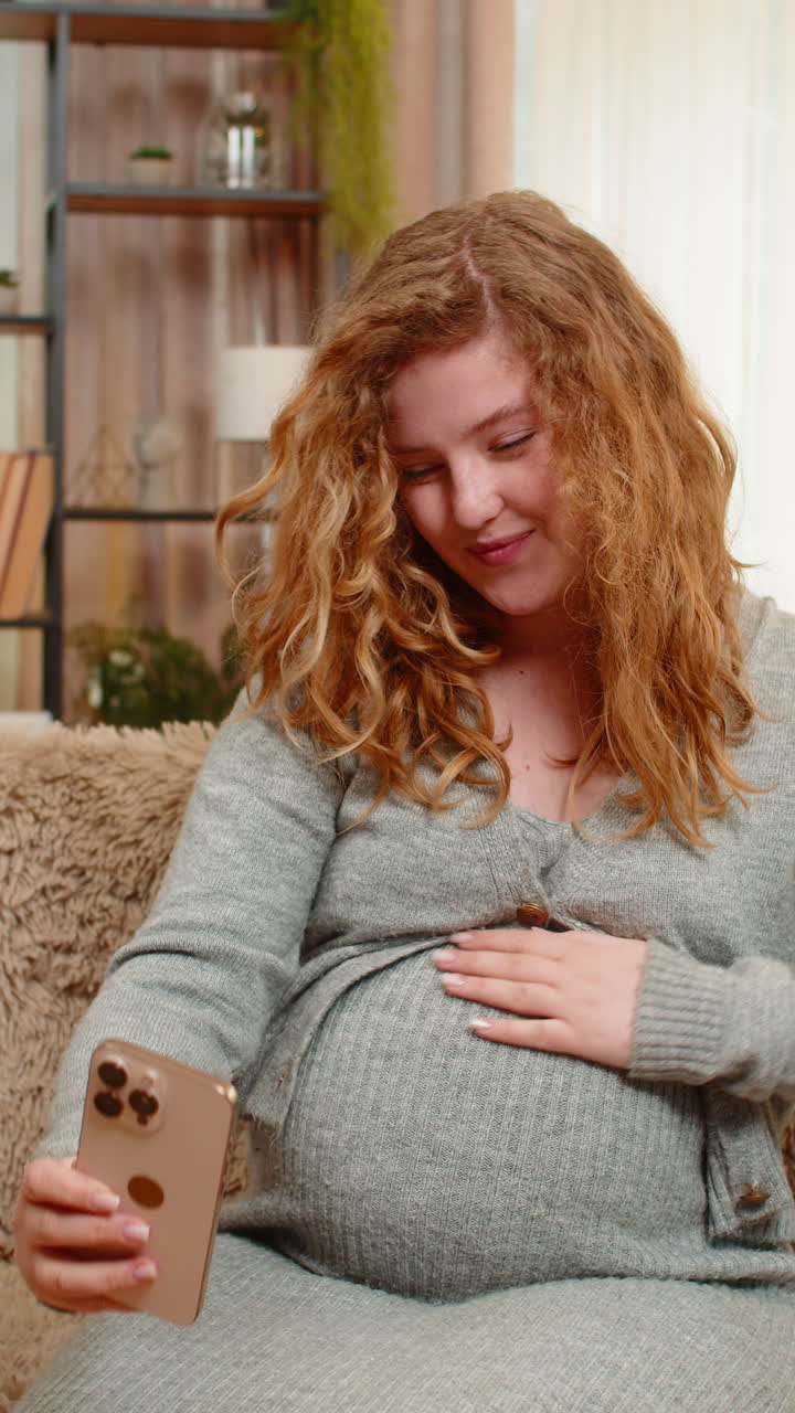 Pregnant young woman talking on video call with friend on smartphone showing belly smiling with joy