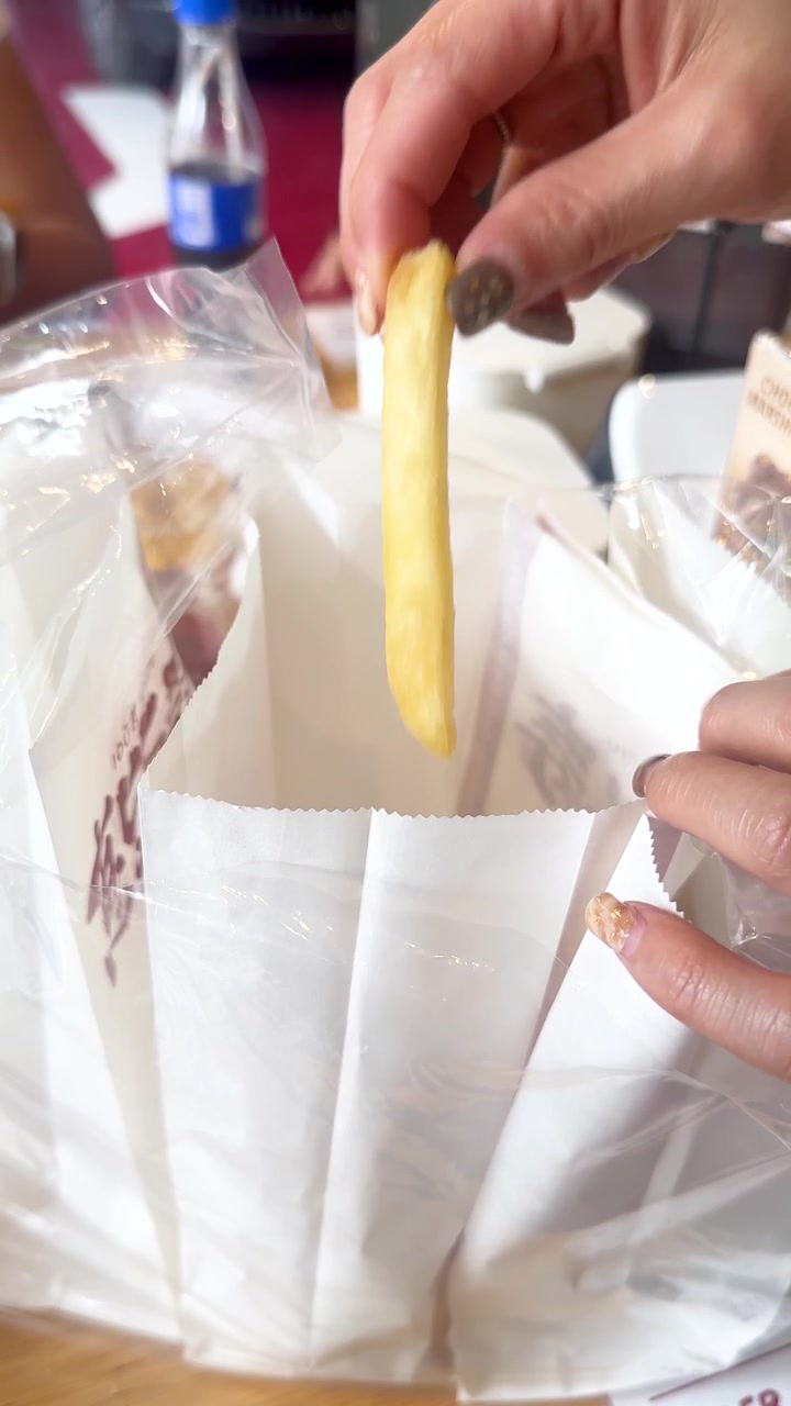 Person retrieves French fry from paper bag at restaurant table, natural lighting, close-up perspective