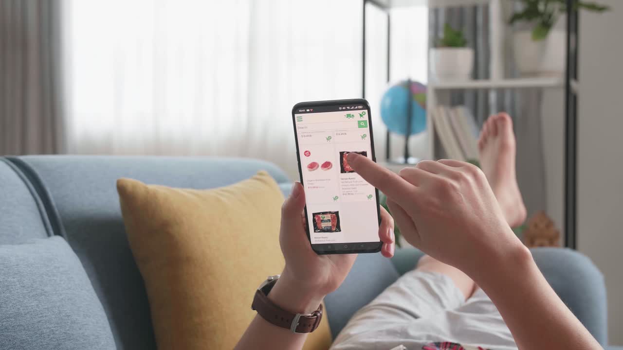Man Lying On Couch Using Smartphone Buys In Internet Shop. Male Selects Beef In Grocery Online Store
