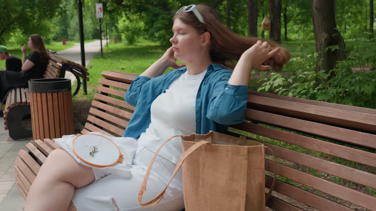 Young lady sitting outdoors on wooden bench in green park drops scissors into fabric bag, adjusts hair, and relaxes on warm sunny day, enjoying peaceful atmosphere surrounded by trees and nature