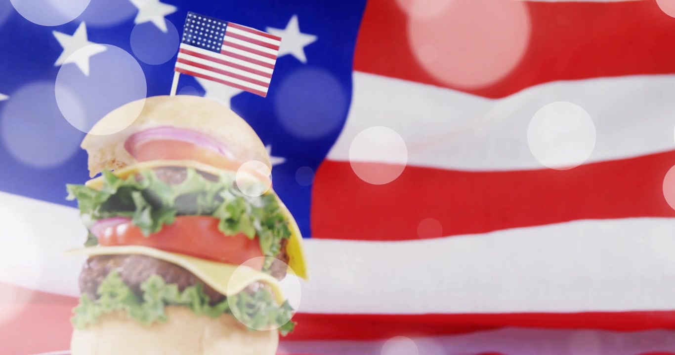 カメラが迫り、積み重ねられたチーズバーガーが上昇し、ミニチュアのアメリカ国旗が中央に配置され、ボケが漂い誇りを呼び起こす。