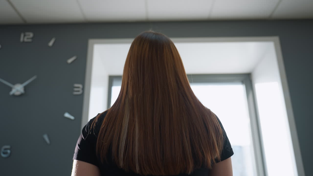 back view of athlete with long straight brown hair facing large window with light streaming through, modern wall clock on dark wall in gym setting, natural light highlighting hair and shoulders