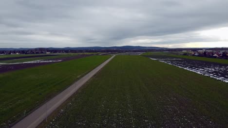 Toma-De-Drones-De-Campos-Marrones-Y-Verdes-En-Invierno-Con-Poca-Nieve-Y-Camino-Recto-En-Dirección-De-Movimiento