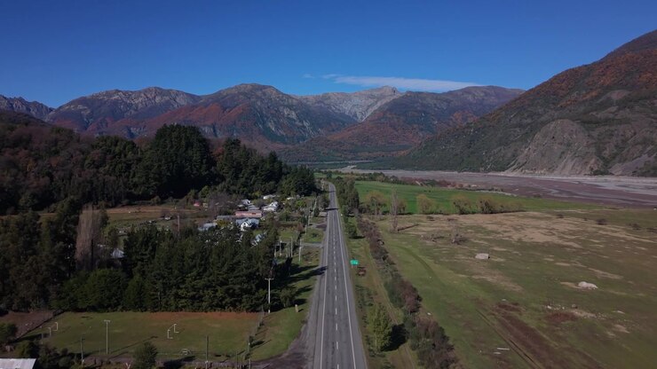 Volo in avanti su un'autostrada rurale oltre una piccola città agricola verso un'ampia valle incorniciata da aspre montagne autunnali dai colori vivaci e un ampio letto di fiume intrecciato