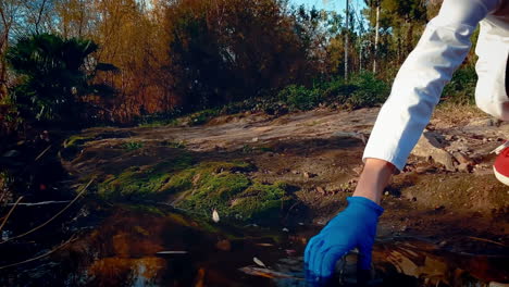 a young woman scientist at a creek, wearing protective eyewear and a lab coat, taking a water sample and holds it up