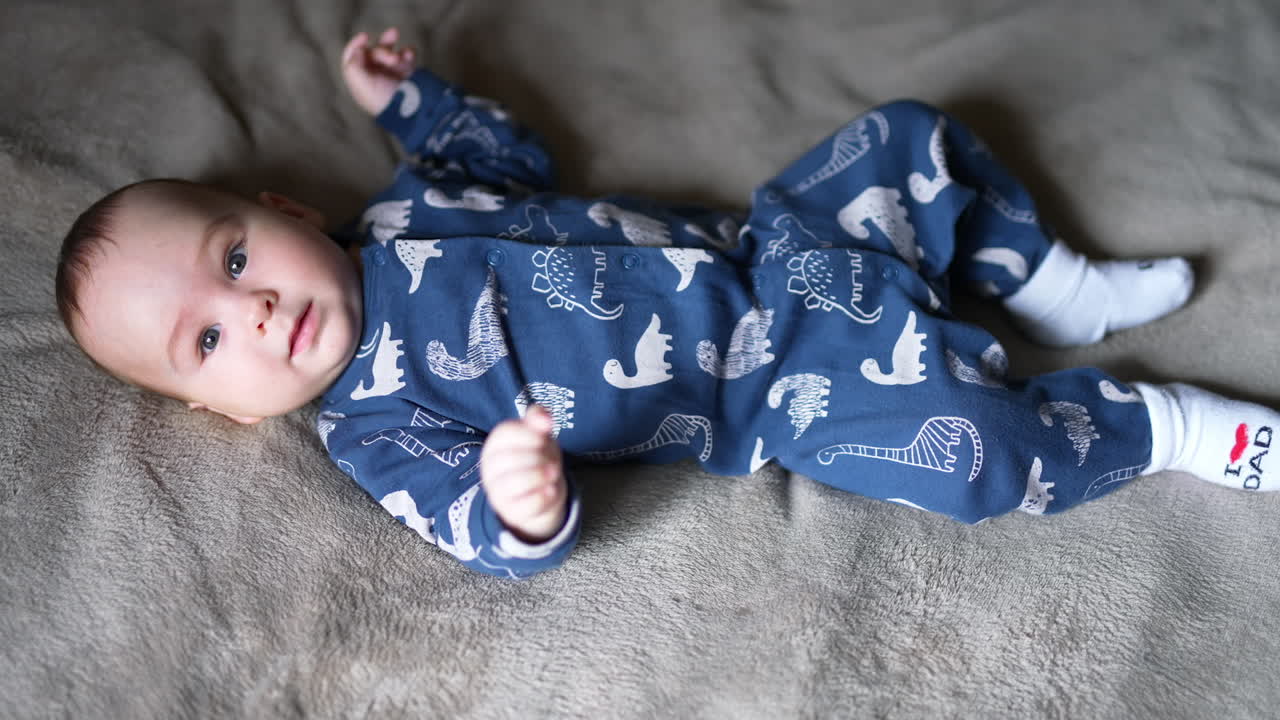 Sweet healthy toddler boy in blue clothes lies on bed. Adorable boy looking into camera and smiling sweetly. Top view.