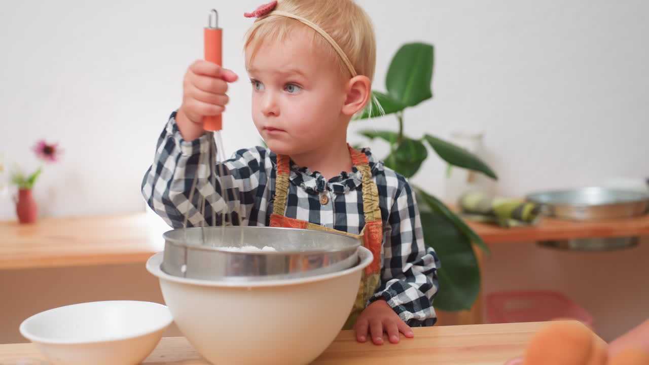 Bambina che indossa un grembiule usa una frusta per setacciare la farina in una ciotola mentre un altro bambino parzialmente visibile osserva nelle vicinanze, con un'espressione concentrata che mostra apprendimento e concentrazione durante la preparazione in cucina