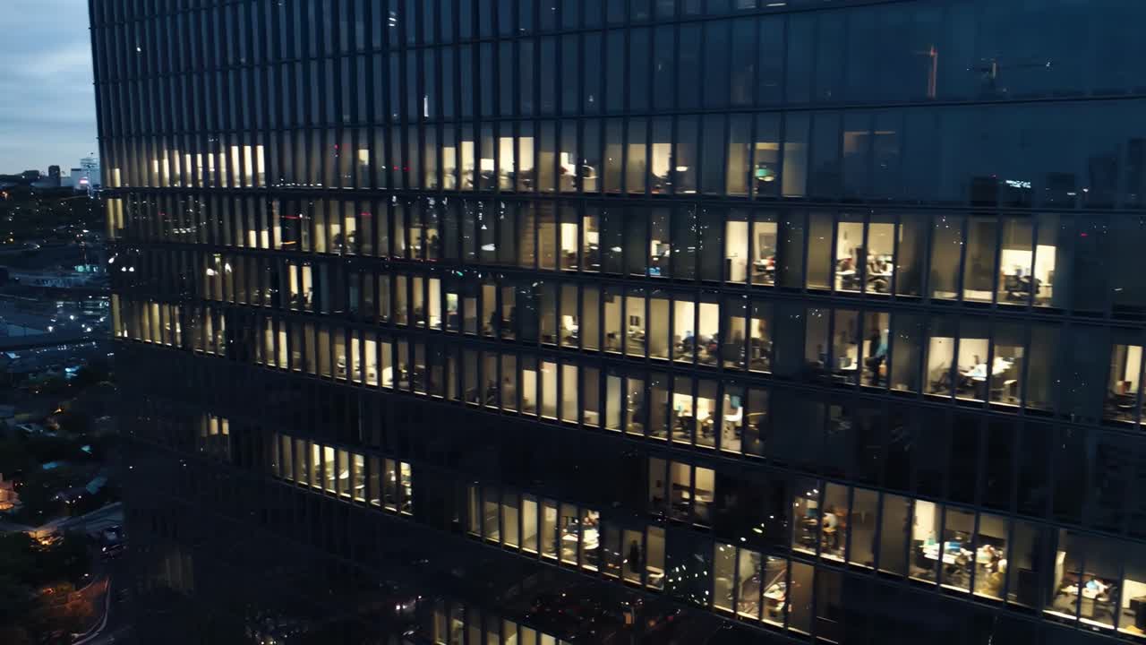 Vídeo aéreo de um prédio de escritórios moderno à noite, mostrando janelas iluminadas e a vida na cidade
