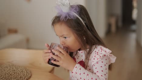 una niña toma un sorbo de un vaso mientras está sentada en una mesa de madera. lleva una camiseta de mangas largas con un patrón de corazón y una cinta para la cabeza, disfrutando de un momento tranquilo en un ambiente cálido y acogedor del hogar.