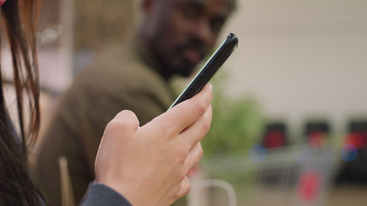 Close up view of woman holding smartphone and typing while seated indoors, with blurred man in background glancing at her with unhappy expression