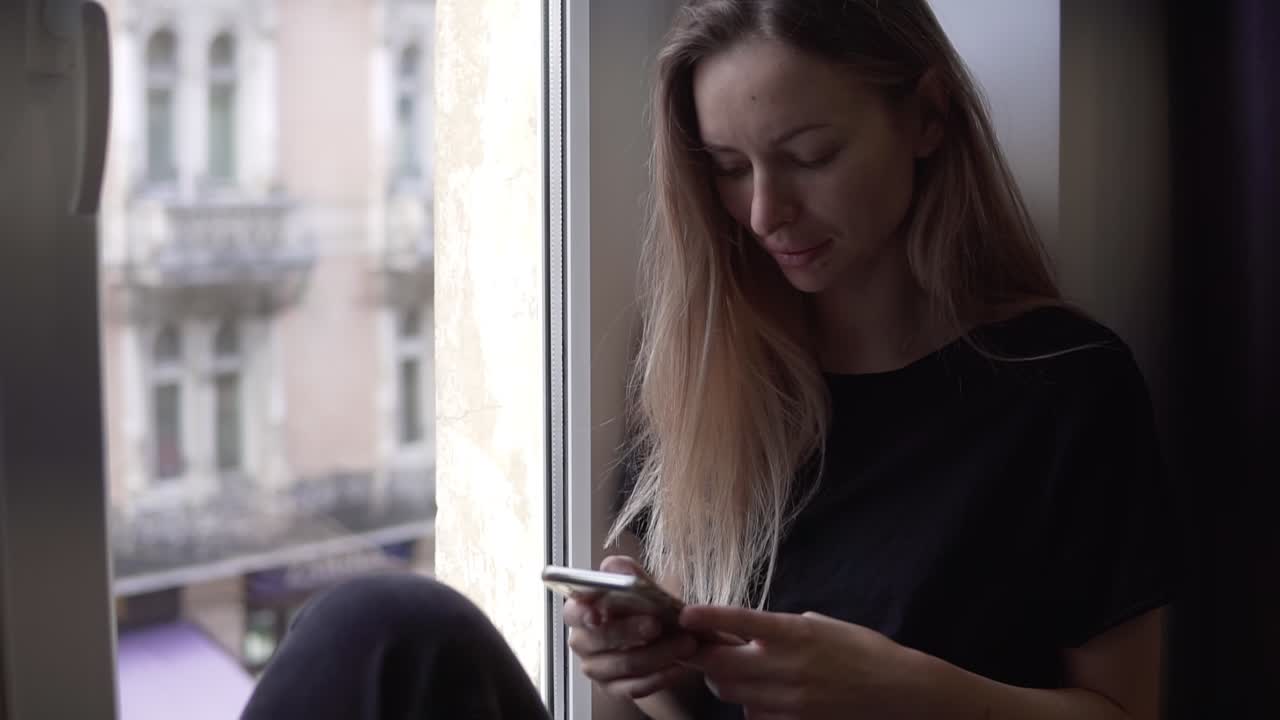 Portrait of a woman sitting on window sill and using smartphone