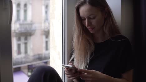 Retrato-De-Una-Mujer-Sentada-En-El-Alféizar-De-La-Ventana-Y-Usando-Un-Teléfono-Inteligente