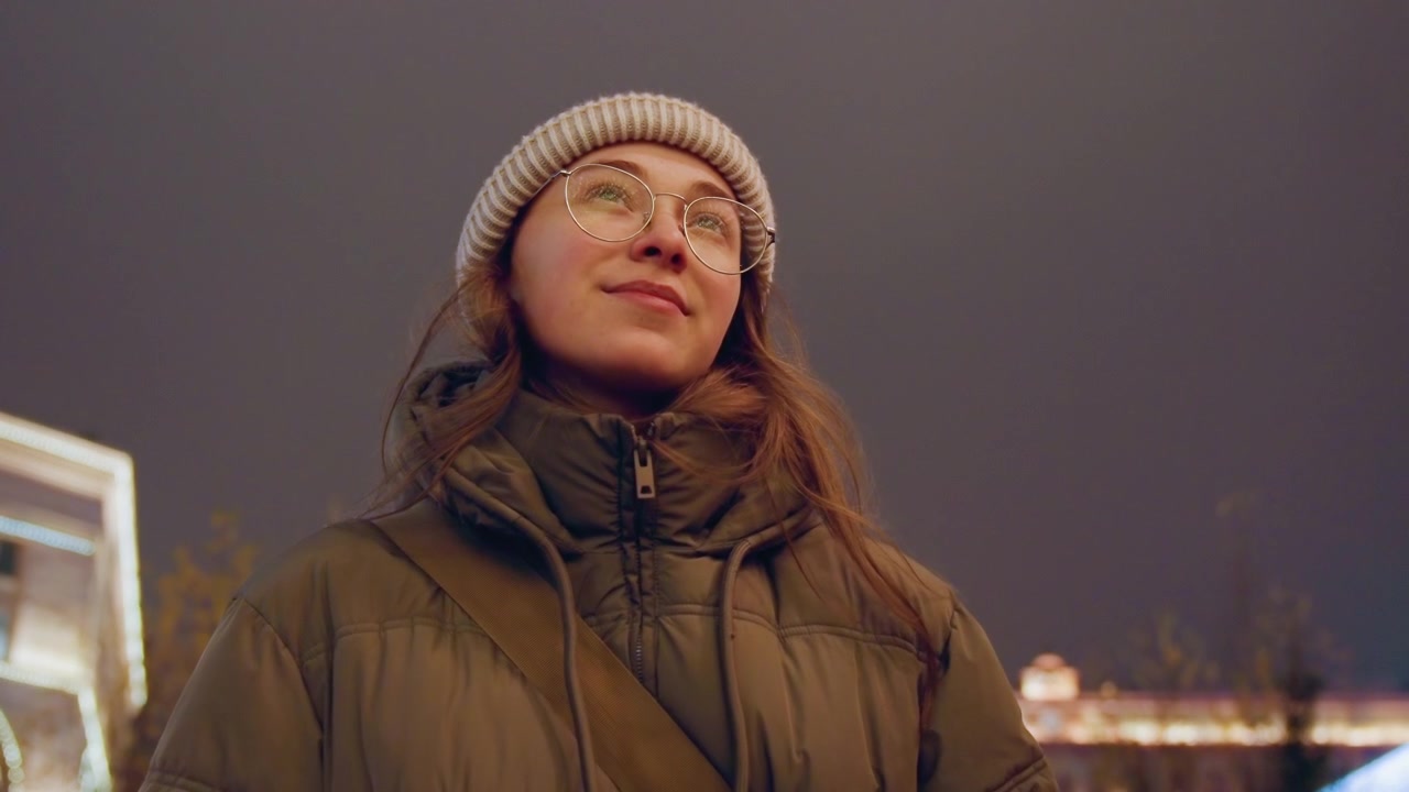 Ritratto notturno urbano di una studentessa bianca che ammira le luci festive, indossando un piumino, un berretto e occhiali; un caldo bagliore dai lampioni e dagli edifici decorati, un sorriso contemplativo, il respiro visibile nel freddo.