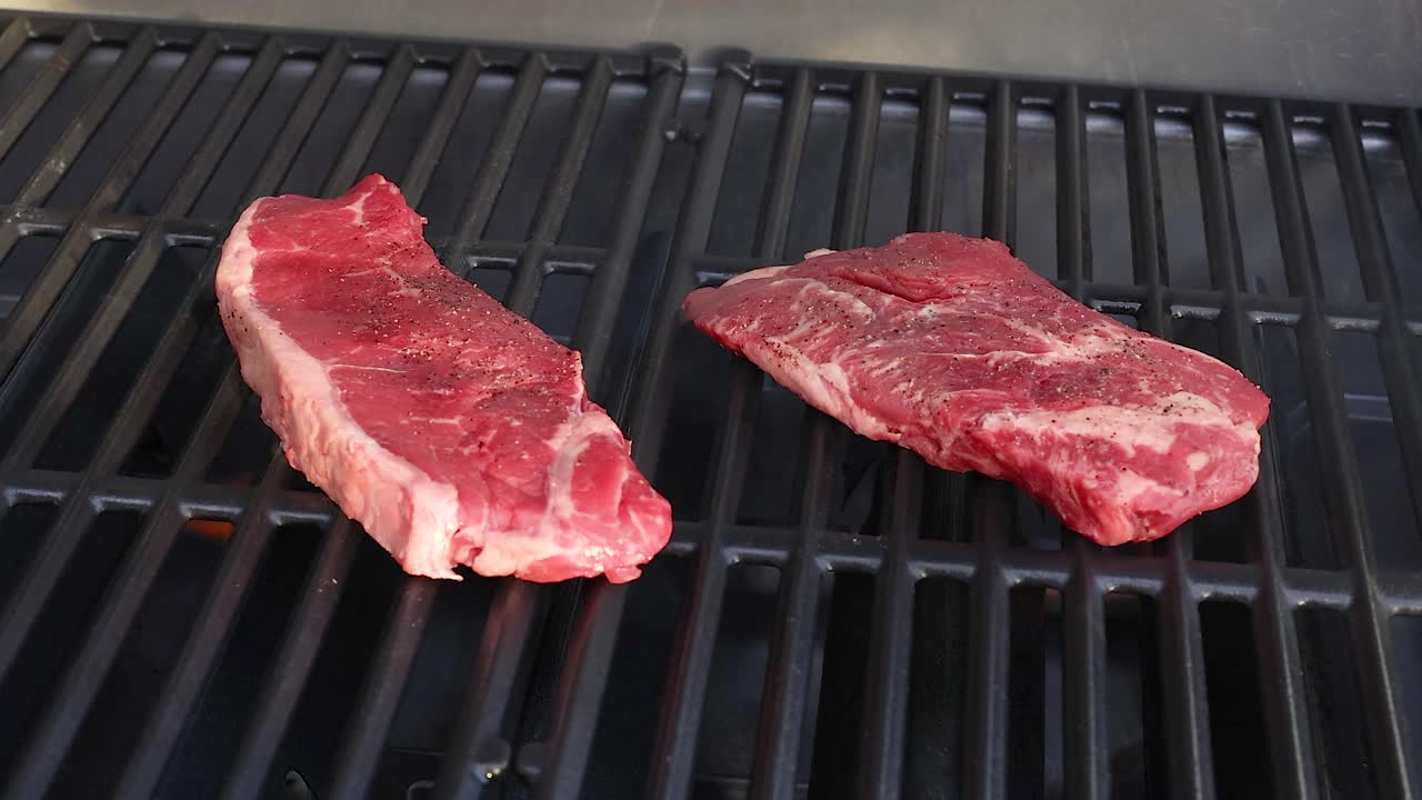 Two raw beef steaks placed on a barbecue grill, gradually cooking over open flames with visible grill marks