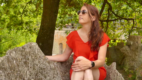 Young-woman-wearing-a-red-dress,-sitting-on-a-bench-in-the-Jardim-das-Virtudes-in-Porto