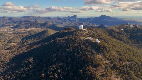 drone footage glides over forested hills toward a large observatory dome at siding spring, under clear daylight with dramatic mountain backdrops