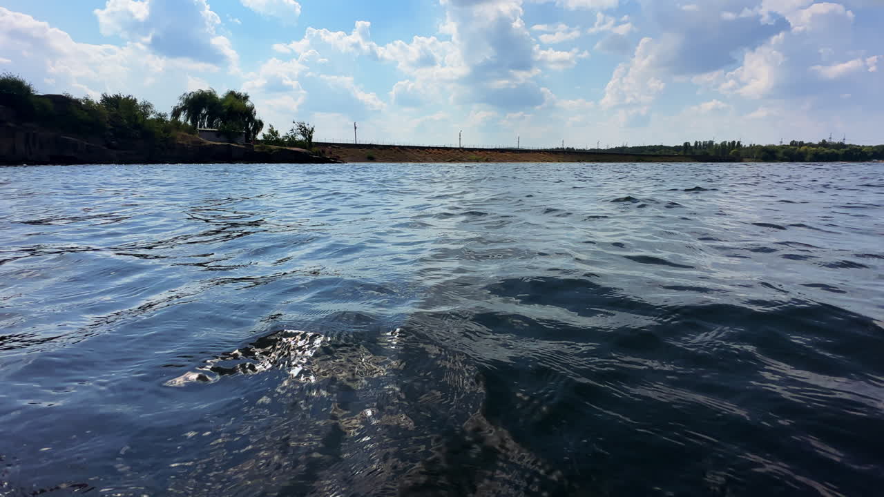 Uneven surface of river. Little waves move in the wind. Close up. Waterfront and sky at backdrop.