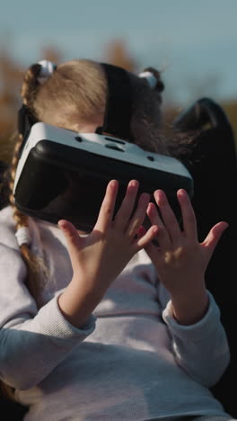 kid in wheelchair plays computer game using vr headset. preschooler girl rests sitting in wheelchair on river bank on blurred background closeup